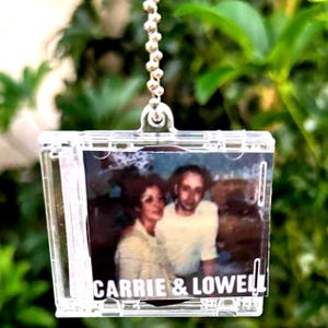 May include: A clear, rectangular keychain with a photo of two people. The photo is inside a clear plastic case and is attached to a silver ball chain. The text "CARRIE & LOWELL" is printed at the bottom of the photo.