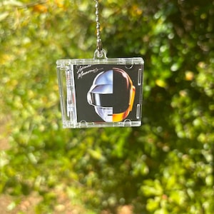 May include: A clear plastic CD case pendant with a silver chain. The CD cover art features a black background with the Daft Punk logo and a helmet design in silver, orange, and yellow. The pendant hangs against a blurred green background.