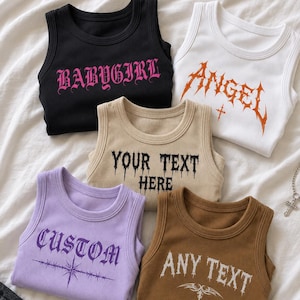 Könnte beinhalten: Fünf gerippte Tanktops in Schwarz, Weiß, Beige, Lavendel und Braun. Das schwarze Oberteil hat den rosa Schriftzug BABY GIRL. Das weiße Oberteil zeigt den orangefarbenen Schriftzug ANGEL. Das beige Oberteil trägt YOUR TEXT HERE, das lavendelfarbene CUSTOM und das braune ANY TEXT.