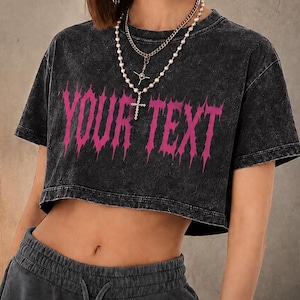 Op de afbeelding: Een zwart acid-wash crop t-shirt met de tekst "YOUR TEXT" in een grillig, felroze lettertype op de borst. Het shirt heeft korte mouwen en een ronde hals. De afbeelding bevat ook de tekst "CUSTOM CROP TEE" en "MADE YOUR WAY".