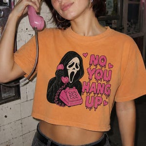 May include: An orange cropped t-shirt featuring a graphic of a black-hooded figure holding a pink telephone. The text reads "NO YOU HANG UP" in pink, dripping letters, accompanied by small pink hearts. The shirt is paired with dark gray pants.