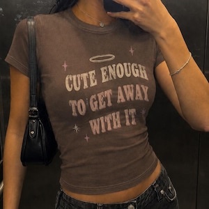 May include: A brown short-sleeved t-shirt featuring the text "CUTE ENOUGH TO GET AWAY WITH IT" in a light-coloured font. The design includes a halo graphic above the text and small star accents on the sides, printed on a fitted cotton fabric.