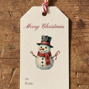 Könnte beinhalten: Ein Weihnachtsgeschenkanhänger mit einer Aquarellillustration eines Schneemanns mit Zylinder und Schal, der eine Zuckerstange hält. Der Anhänger trägt den roten Schriftzug "Merry Christmas" mit Platz für "an:" und "von:" auf einem Holzgrund.