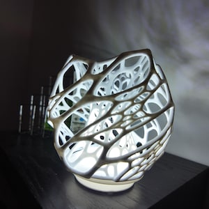 Op de afbeelding: Een witte, open geweven decoratieve lamp met een uniek, organisch ontwerp. De lamp heeft een afgeronde vorm met een netwerk van onderling verbonden, webachtige structuren. De lamp wordt van binnenuit verlicht, waardoor een zachte gloed ontstaat.