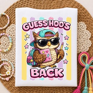 May include: A white t-shirt featuring a graphic of an owl wearing a teal baseball cap and black sunglasses. The owl holds a pink notebook and a yellow pencil, with a teal backpack. The text reads GUESS HOO'S BACK in pink bubble letters.