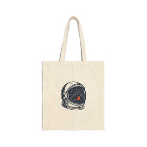Peut inclure: Sac fourre-tout crème avec une illustration détaillée d'un casque d'astronaute. Le design du casque comprend des étoiles, des planètes et un croissant de lune orange et blanc. Le sac a un design simple avec de longues anses.