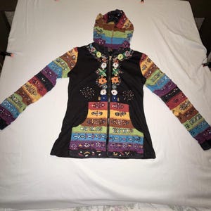 Pode incluir: Casaco com capuz preto com zíper e mangas e capuz estampados coloridos. A frente apresenta desenhos florais bordados e dois bolsos com listras horizontais de várias cores. As mangas e o capuz têm um design de patchwork.