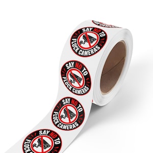May include: A roll of circular adhesive stickers featuring a black and white graphic of a surveillance camera inside a red prohibition circle. The text reads SAY NO TO FLOCK CAMERAS in bold black and red lettering against a white background.