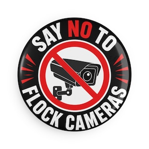 Puede incluir: Una chapa redonda con fondo negro que muestra una cámara de seguridad tachada por una línea diagonal roja. El texto SAY NO TO FLOCK CAMERAS aparece en letras mayúsculas de color blanco y rojo alrededor del diseño central.