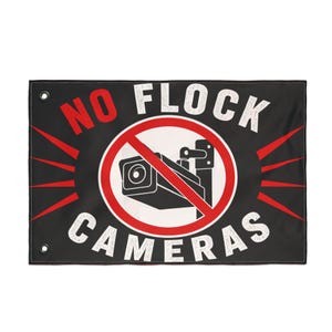 May include: A rectangular black flag featuring the red text NO and white text FLOCK CAMERAS. A central white circle contains a black security camera icon crossed out by a red diagonal line. Red rays extend outward from the circle, and two metal grommets are visible on the left side.