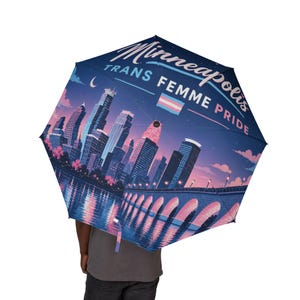 Op de afbeelding: Een donkerblauwe paraplu met een grafische illustratie van de skyline van Minneapolis bij nacht met een brug over het water. De tekst Minneapolis TRANS FEMME PRIDE is gedrukt in wit en roze, vergezeld van een kleine transgender vlag.