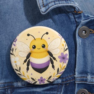 Könnte beinhalten: Ein runder Ansteckbutton mit einer zentralen Illustration einer flauschigen Hummel mit gelben, weißen und violetten Streifen. Die Hummel ist von einem dekorativen Kranz aus kleinen violetten und weißen Blüten sowie schwarzen Blättern umgeben.