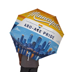Op de afbeelding: Een paraplu met een afbeelding van de skyline van Minneapolis bij nacht met een reflectie in het water. Het ontwerp bevat de tekst "Minneapolis" en "ARO-ACE PRIDE" in donkerblauwe letters, samen met een klein hartje in oranje, wit en blauw.