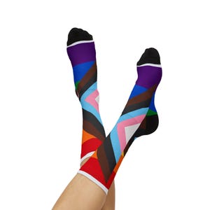 Op de afbeelding: Een paar kniehoge sokken met een kleurrijk geometrisch ontwerp. De sokken hebben een zwarte teen en hiel, met een witte band aan de bovenkant. Het ontwerp bevat verschillende kleuren, waaronder rood, oranje, bruin, blauw en roze, gerangschikt in een diagonaal patroon.