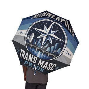 Op de afbeelding: Een marineblauwe en lichtblauwe paraplu met een afbeelding van de skyline van Minneapolis, een brug en een grote kompasster. De tekst luidt MINNEAPOLIS en TRANS MASC PRIDE in witte en lichtblauwe letters tegen een geometrische achtergrond.