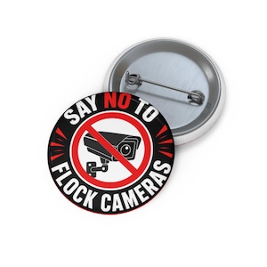 May include: A round pin-back button featuring a black and white graphic of a surveillance camera inside a red prohibition circle. The text reads SAY NO TO FLOCK CAMERAS in bold white and red lettering against a black background with red accents.