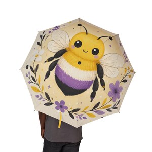 May include: A cream-colored umbrella featuring a large, central illustration of a fuzzy bee with yellow, white, and purple stripes. The bee is surrounded by small purple and white flowers, black leafy stems, and tiny star accents on a light background.