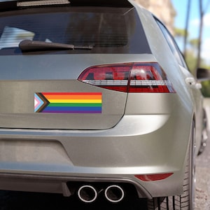 May include: A rectangular bumper sticker featuring the Progress Pride flag design, which includes the traditional six-stripe rainbow flag combined with a chevron pattern of light blue, pink, white, brown, and black stripes, applied to a car.