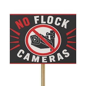 May include: A rectangular sign with a black background and red border featuring the white text NO FLOCK CAMERAS. A central white circle contains a black security camera icon crossed out by a red diagonal line. The sign is mounted on a wooden stake.