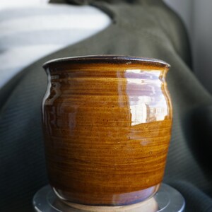May include: A handcrafted, brown ceramic jar with a glossy finish. The jar has a wide, rounded body and a slightly flared rim. The surface features horizontal striations, adding texture. The jar sits on a silver metal stand, with a dark green fabric in the background.