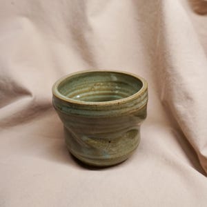 May include: Handmade ceramic cup with a unique, textured design. The cup features a light green and brown color palette, with visible ridges around the rim. The cup's form is slightly asymmetrical, adding to its handcrafted appeal. A rustic, earthy piece.