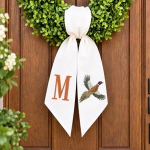 May include: A white fabric ribbon hanging from a green wreath. The left side of the ribbon features a large, embroidered orange letter M, while the right side displays an embroidered, multi-colored pheasant in flight.