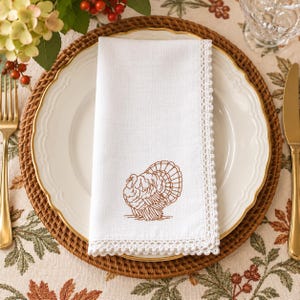 May include: A white cloth napkin featuring a brown embroidered turkey design on the lower section and a delicate white crocheted lace border along the edges. The napkin is folded and placed centrally on a white dinner plate with a gold rim.