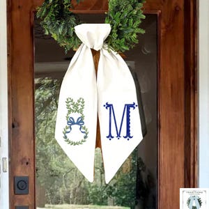 May include: A white fabric door hanger with two long, pointed tails hanging from a green wreath. The left tail features an embroidered green leafy bunny silhouette with a blue bow, and the right tail displays a large, dark blue embroidered letter M.