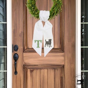 May include: White door sash with a green letter "T" and a black dog illustration. The sash is hanging on a wooden door with a green wreath. The door has a black handle and lock.