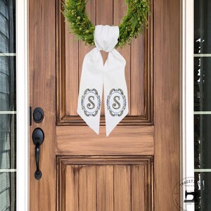 May include: A white door sash with the letter "S" in a decorative oval design, hanging from a green wreath. The sash is tied at the top and hangs vertically. The door is made of wood.