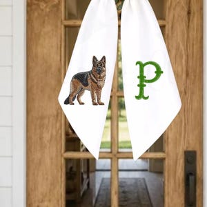 May include: A white fabric door hanger bow featuring a detailed embroidered German Shepherd dog on the left tail and a large, textured green embroidered letter P on the right tail, suspended from a green wreath on a wooden door.