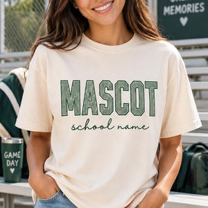 May include: A cream-colored short-sleeve t-shirt featuring the word MASCOT in large, dark green, textured block letters, with the words school name written in a smaller, cursive dark green font centered directly underneath the main text.