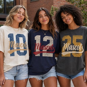 May include: Three short-sleeved t-shirts featuring large distressed numbers and the word Mascot in script. The left shirt is cream with blue 10, the center is navy with white 12, and the right is charcoal with gold 25. All shirts have a crew neck.