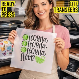 May include: A woman holds a DTF transfer sheet with the words "Tequila Floor" in black script, accented with green circles and a lime slice. The sheet is labeled "Ready to Press" and "DTF Transfers" in yellow.