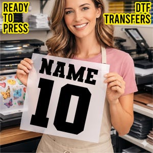 May include: A clear DTF transfer sheet featuring the black text NAME and the number 10 in a bold athletic font. The text READY TO PRESS DTF TRANSFERS is visible in the corners. The sheet is held up, displaying the design clearly for application.