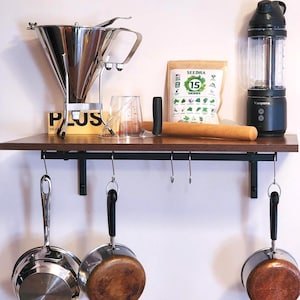 May include: A kitchen shelf displays various cooking tools and ingredients. A stainless steel funnel, measuring cup, and a bag of herbs are visible. A blender and rolling pin are also present. Hanging below are stainless steel pans.