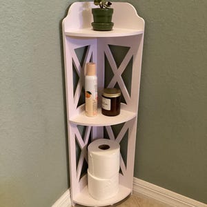 May include: A white corner shelf unit with three tiers. The top shelf holds a small potted plant. The middle shelf has a bottle and a jar. The bottom shelf holds two rolls of white toilet paper. The unit is in a bathroom.