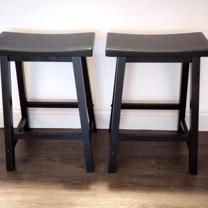May include: Two black wooden bar stools with curved seats. The stools have a simple, modern design with four legs and a footrest. The stools are placed in front of a white wall and a dark wood floor.