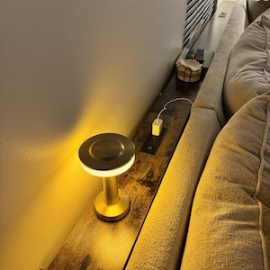 May include: A small, gold-colored bedside lamp with a round, illuminated top sits on a wooden shelf. The shelf is mounted between a wall and a sofa. A charging port and a charging cable are also visible.