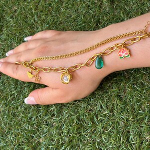 Peut inclure: Un bracelet doré à double chaîne composé d'une chaîne à maillons ovales épais et d'une chaîne plus fine. Le bracelet est orné de plusieurs breloques, dont une goutte verte, une fleur de lotus rose et verte, et de petites formes dorées.