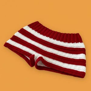 以下が含まれることがあります： 赤と白のストライプ柄のかぎ針編みショートパンツ。ショートパンツは、赤と白の水平ストライプが特徴です。ウエストバンドとエッジは無地の赤色です。オレンジ色の背景に表示されています。