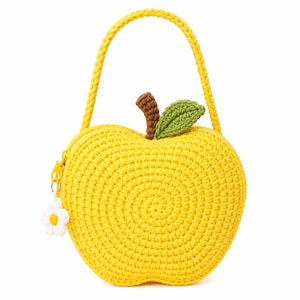 May include: A bright yellow apple-shaped crochet handbag featuring a single braided handle, a brown stem, and a green leaf. The bag includes a yellow zipper closure with a small white and yellow flower charm attached to the zipper pull.
