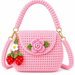 Puede incluir: Un bolso de ganchillo rosa claro con asa superior y correa de hombro desmontable con herrajes dorados. La solapa frontal está decorada con una flor rosa tridimensional con hojas verdes y un colgante que representa dos fresas de color rojo.