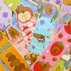 May include: A collection of colorful bookmarks featuring cartoon capybaras in various designs. The bookmarks showcase capybaras with apples, strawberries, and in a seashell, with a variety of pastel backgrounds and patterns.