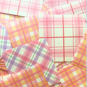 May include: A collection of small, square cards with a plaid pattern in shades of pink, peach, and white. The cards are scattered, showing various angles of the plaid design. The overall theme is stationery or paper goods.