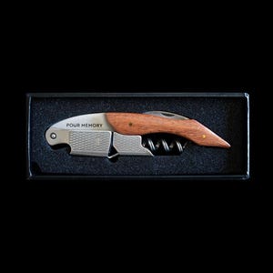 May include: A waiter's corkscrew with a wooden handle and a stainless steel body featuring a textured grip and a spiral worm. The metal surface is engraved with the text "POUR MEMORY". The tool is presented inside a black foam-lined box.