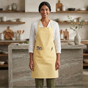 May include: A yellow apron with a geometric pattern. The apron has a large front pocket with measuring spoons and a pen. The apron is worn in a kitchen setting.