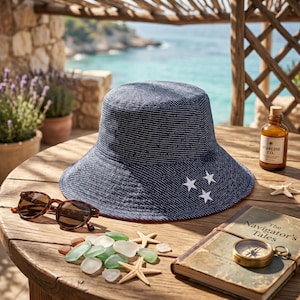May include: A navy blue bucket hat with white star accents and a red brim sits on a wooden table. Sunglasses, sea glass, starfish, a compass, and a bottle with the text "STARLINE CO." are also on the table.