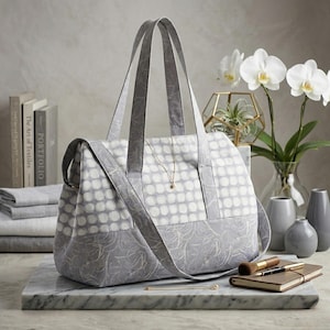 Puede incluir: Un bolso tote gris y blanco con un estampado de lunares en la parte superior y un diseño floral en la parte inferior. El bolso tiene dos asas y una correa para el hombro. Un collar de oro cuelga del bolso. El bolso está sobre una superficie de mármol.