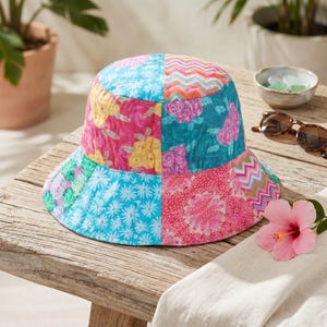 Könnte beinhalten: Ein Patchwork-Eimerhut mit lebendigen floralen, Zickzack- und geometrischen Mustern in Rosa, Blau, Gelb und Grün. Der Hut ist aus gestepptem Stoff gefertigt und liegt auf einer Holzoberfläche.